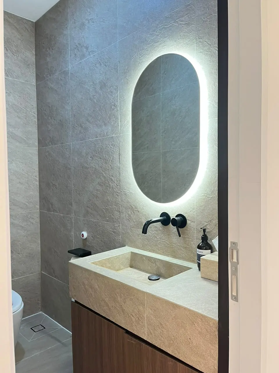 Restroom Hestia Women Clinic dengan vanity batu minimalis, wall-mounted faucet hitam, dinding bertekstur batu, dan cermin oval dengan backlight.