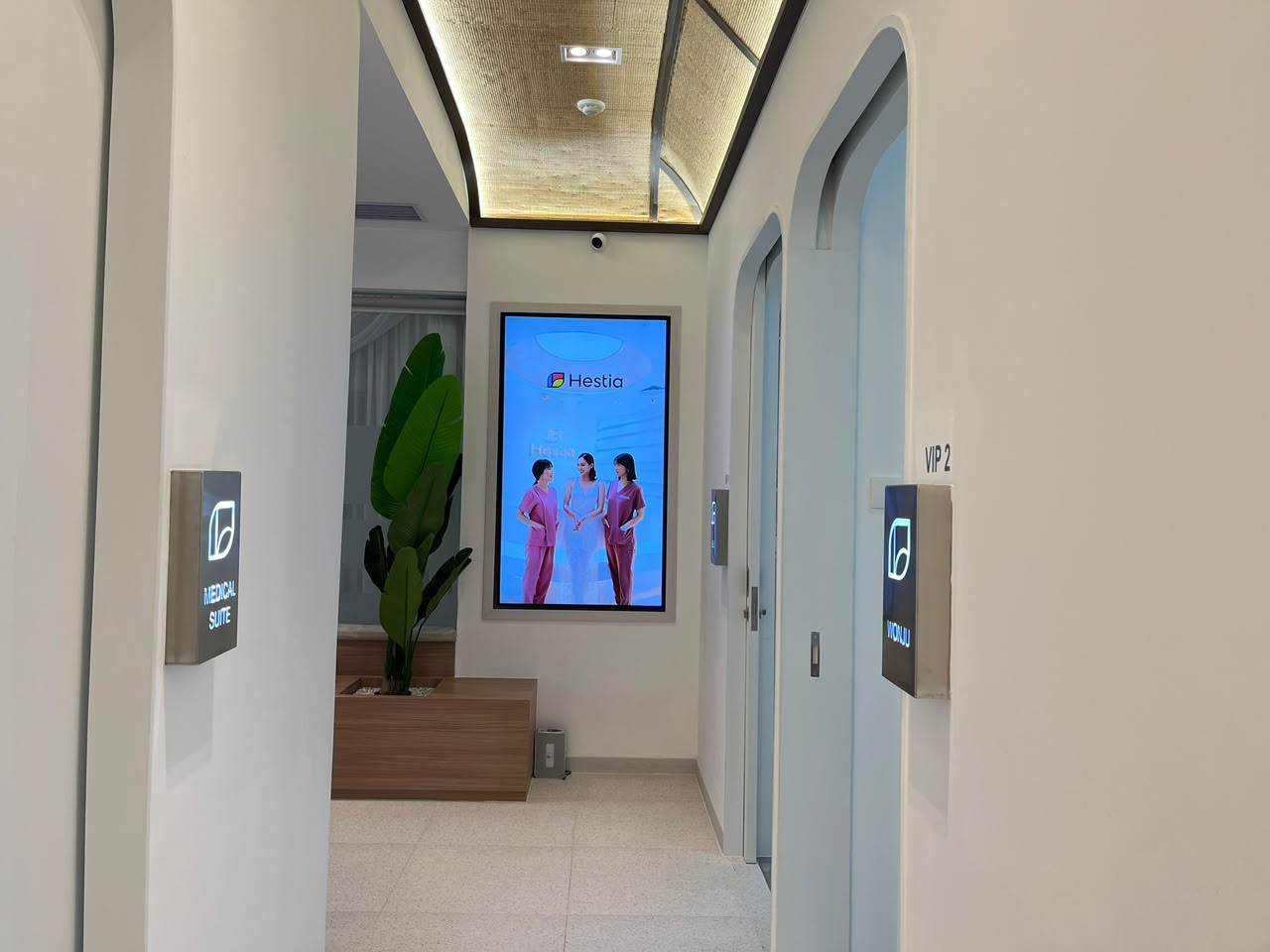 Koridor menuju ruang VIP Hestia Women Clinic dengan signage terintegrasi, bukaan pintu melengkung, planter, ceiling dekoratif, dan digital display.